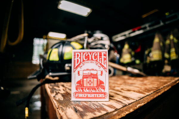Bicycle Firefighters Playing Cards Deck on a wooden table inside of a fire station. Fire gear can be seen blurred in the background.